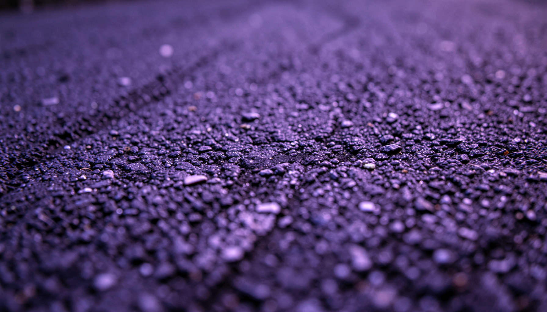 Textura de asfalto escuro com marcas de pneus e pequenas pedras soltas, tons de cinza e preto, iluminação lateral rasante.