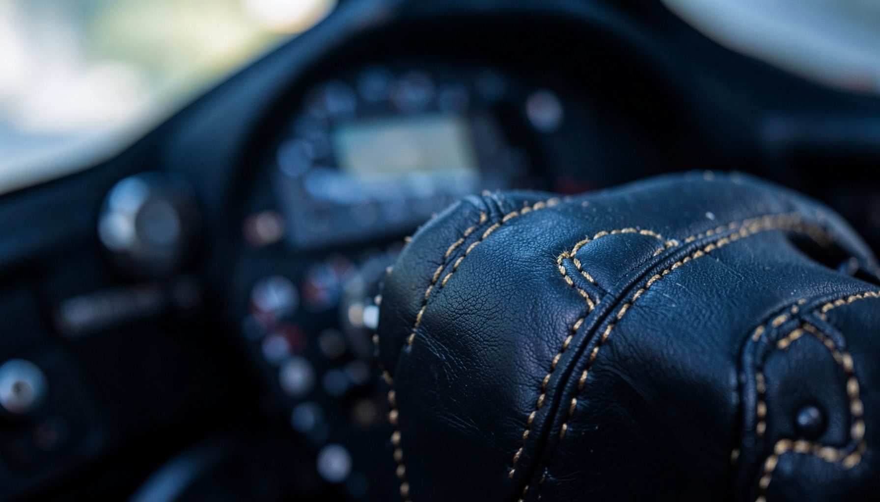 Volante de competição em alcantara preta com costuras douradas, visto de perto contra um fundo desfocado de cockpit.