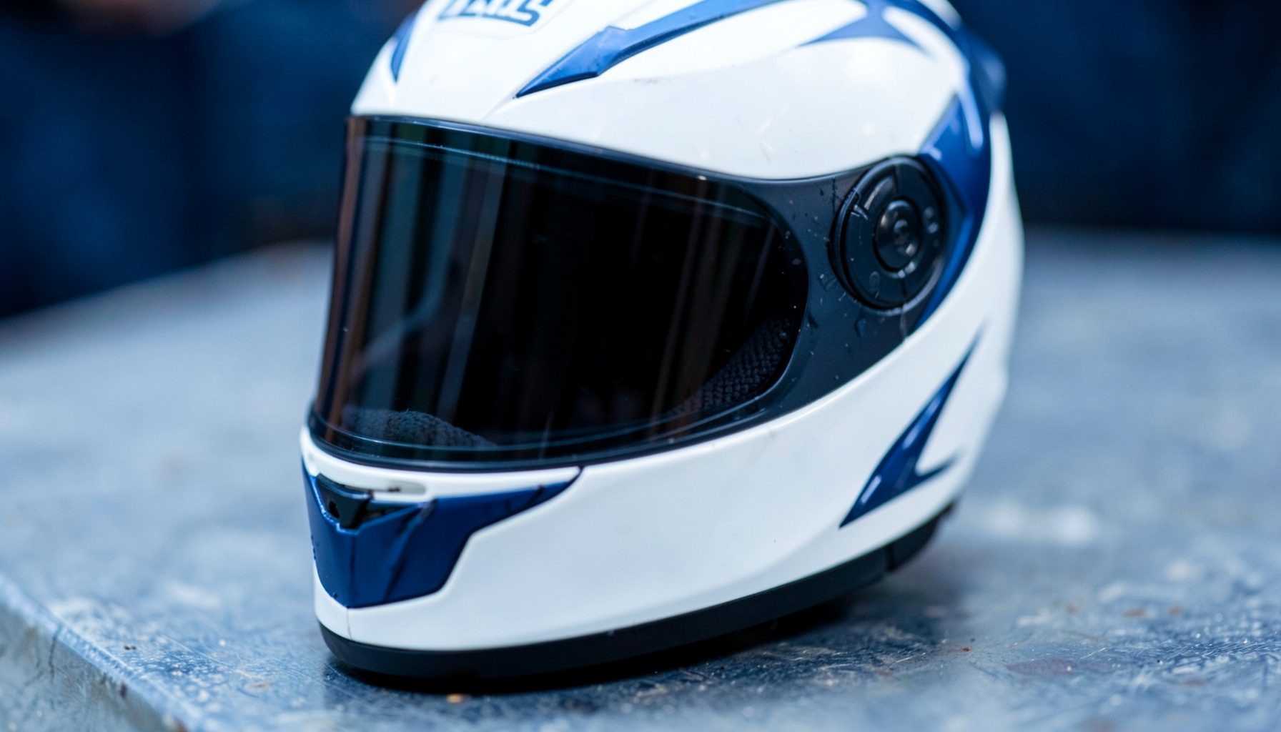 Close-up de capacete de corrida branco com viseira fumada e detalhes em azul-escuro, pousado sobre uma superfície metálica.