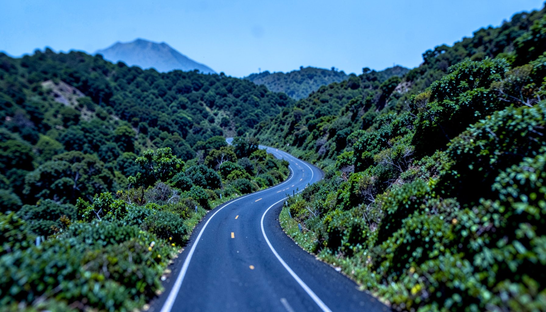 Vista aérea de uma estrada de montanha sinuosa com asfalto escuro, rodeada por vegetação densa em tons de verde profundo, sob um céu azul limpo.