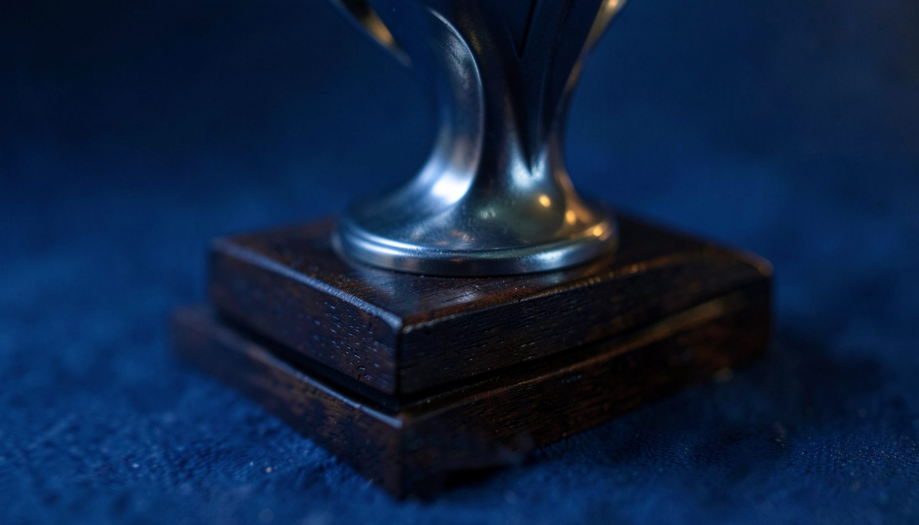 Troféu douado sobre uma base de madeira escura, iluminado por uma luz suave lateral que realça os seus contornos polidos.